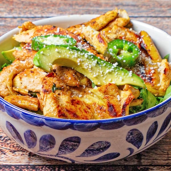 Grilled chicken, avocado slices, and jalapeños in a decorative bowl on a wooden table.