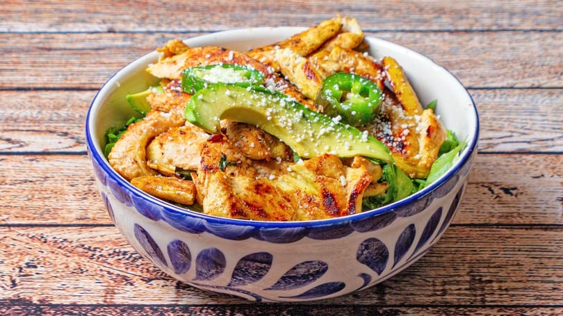 Grilled chicken, avocado slices, and jalapeños in a decorative bowl on a wooden table.