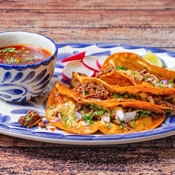 Three birria tacos with consome, radishes, and lime wedges on a blue and white plate.
