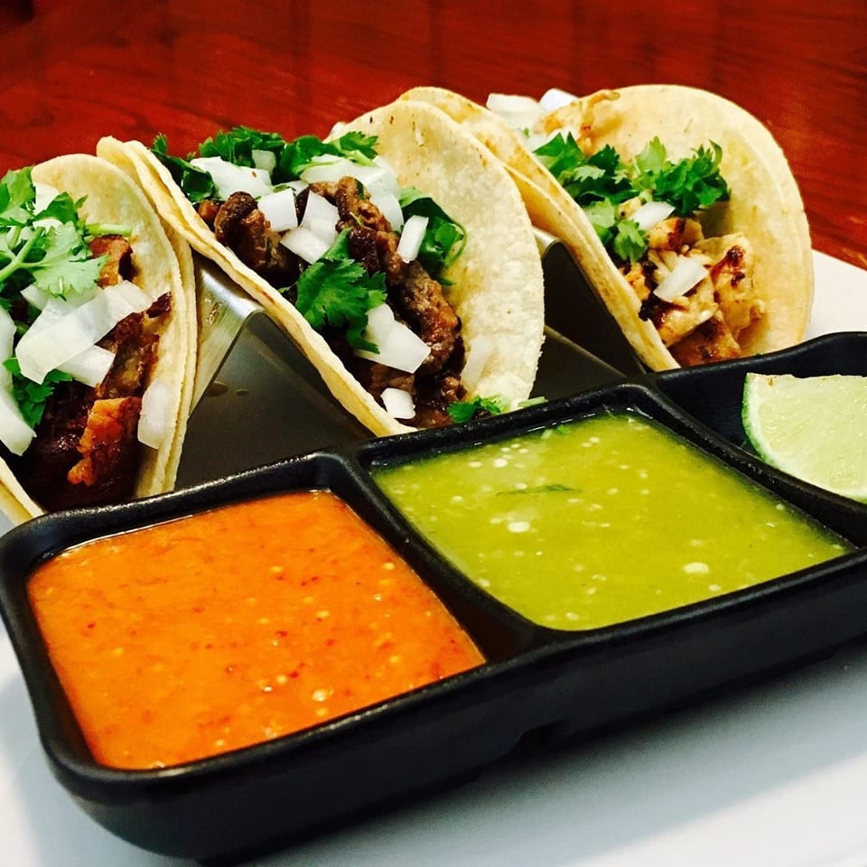 Four tacos with meat, onions, and cilantro, served with red and green salsa and a lime wedge.