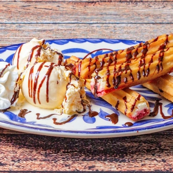 Churros drizzled with chocolate, vanilla ice cream, and whipped cream on a blue and white plate.