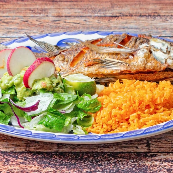 Fried whole fish with rice, guacamole, salad, radishes, and lime on a blue and white plate.