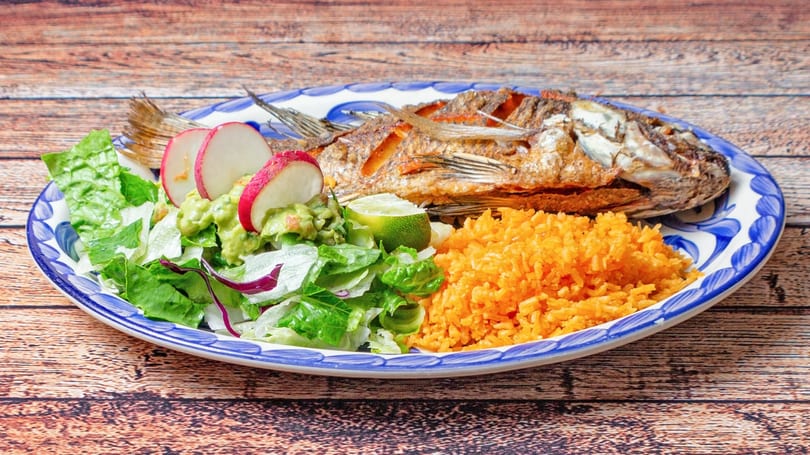 Fried whole fish with rice, guacamole, salad, radishes, and lime on a blue and white plate.