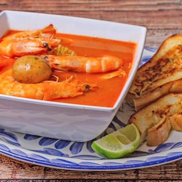 Shrimp soup with potatoes and corn in a white bowl, served with toasted bread and lime.