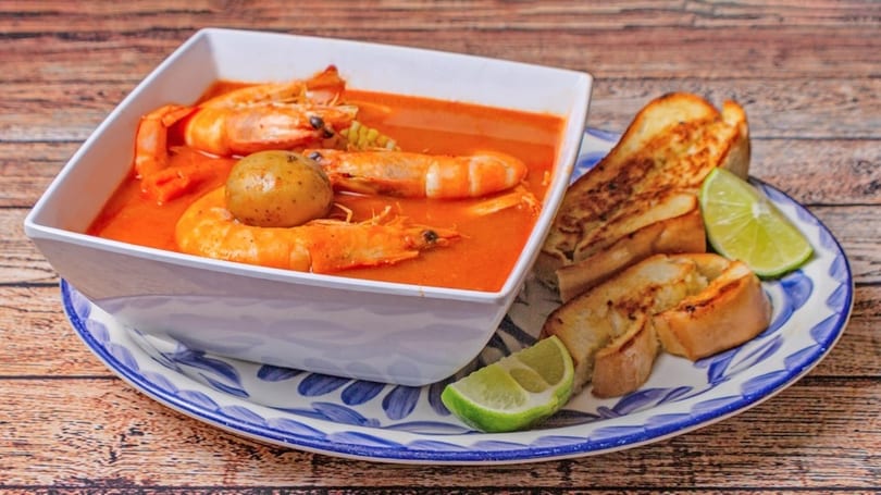 Shrimp soup with potatoes and corn in a white bowl, served with toasted bread and lime.